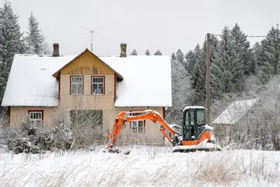 Orange grävmaskin framför ett gult trähus i snön