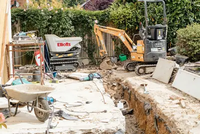 Minigrävare och dumper vid en uppgrävd husgrund i en trädgård