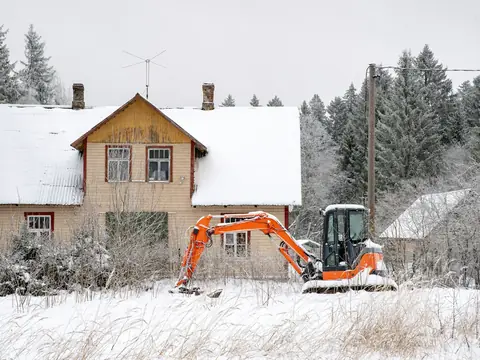 Orange grävmaskin framför ett trähus i snön
